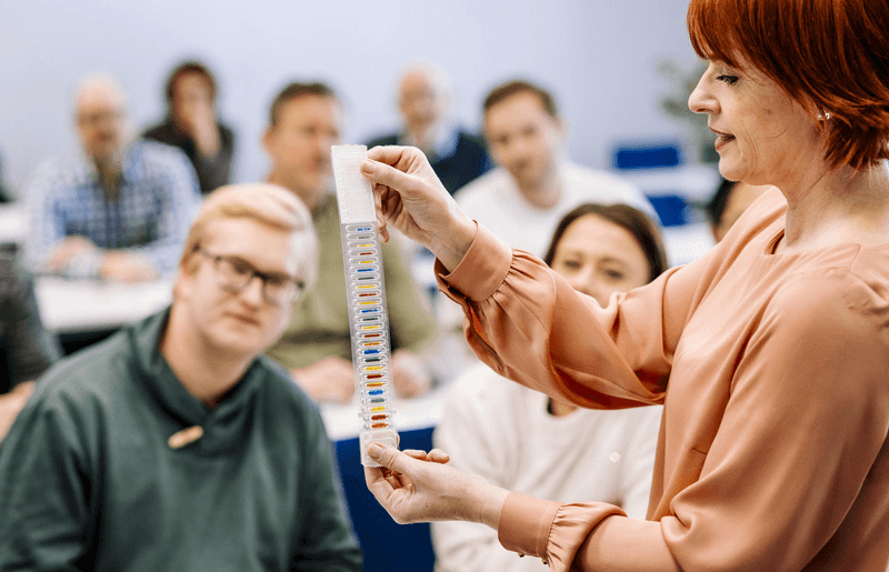Dozentin erklärt im Seminar zum organisatorischen Brandschutz die Funktion farbiger Glasfässchen an Sprinkleranlagen als temperaturempfindliche Auslöseelemente zur automatischen Brandbekämpfung; im Hintergrund folgen Teilnehmerinnen und Teilnehmer aufmerksam der Schulung.